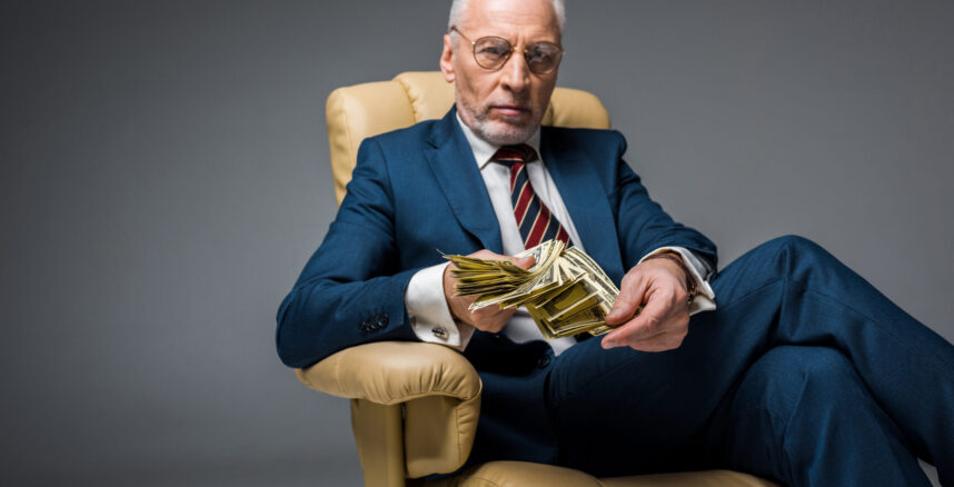 serious mature businessman sitting in armchair and holding dollar banknotes on grey Alem da Fortuna A Importancia da Psicanalise Financeira para Bilionarios 1 scaled