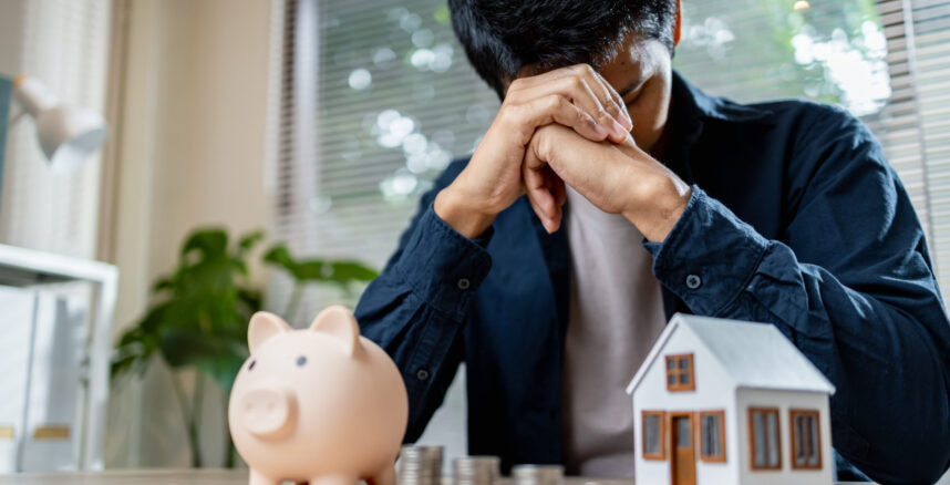worried man sits at table with piggy bank and house model pedro braggio A Armadilha da Casa Propria Quando o Sonho Vira Pesadelo Financeiro scaled