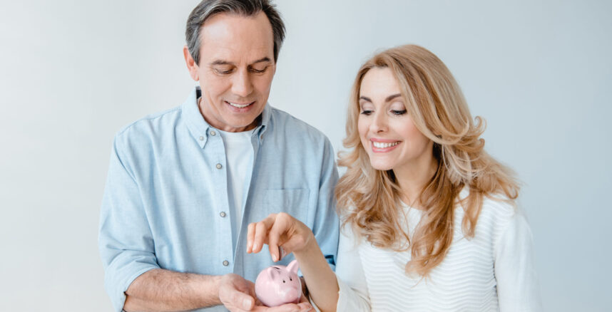 Smiling middle aged couple putting coin into small piggy bank  isolated on grey pedro braggio financas a dois scaled